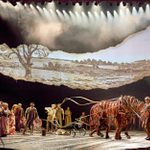 Actors perform on a theater stage featuring a brown horse puppet, controlled by three puppeteers, pulling a plough. The backdrop displays a torn, sepia-toned landscape illustration. Multiple performers, dressed in period costumes, interact and move across the stage amidst dramatic lighting.