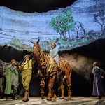 A theatrical scene featuring actors and two life-size horse puppets on stage. One actor rides one of the puppet horses while others stand nearby, one playing an accordion. The background displays an illustration of a rural landscape with trees and buildings.