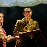 A woman wearing a floral blouse and waistcoat holds a large open book, while a man in a military uniform writes in it. They are on a stage with a dark background, illuminated by a spotlight. The scene suggests a dramatic moment in a theatrical performance.