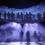 Silhouetted figures of soldiers with rifles charging in formation on a stage. Above them, a backdrop displays an image of more soldiers charging through barbed wire. Bright lights shine down through smoke, creating a dramatic, intense atmosphere.