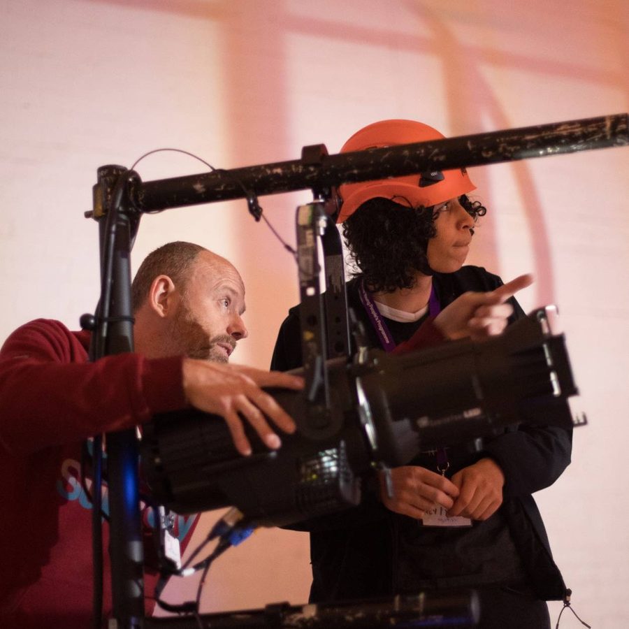 A young person wearing an orange hard hat is shown how to position a theatre light by a technician