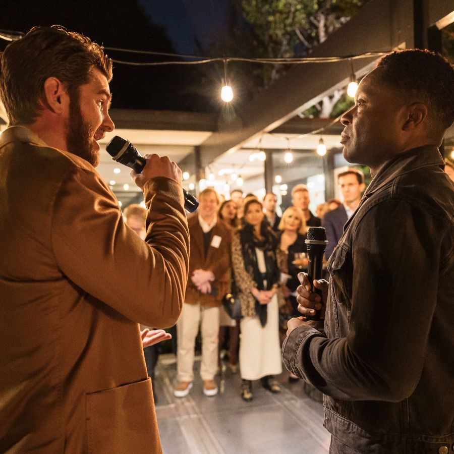 Andrew Garfield speaking into a microphone, standing next to David Oyelowo, with an audience standing in the background.