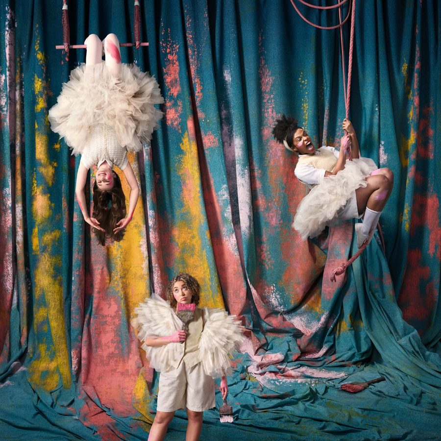 Three performers in whimsical costumes pose against a colorful, painted backdrop. One is upside down on a trapeze, another stands holding a prop, and the third hangs from aerial hoops. They wear fluffy dresses and expressive accessories.