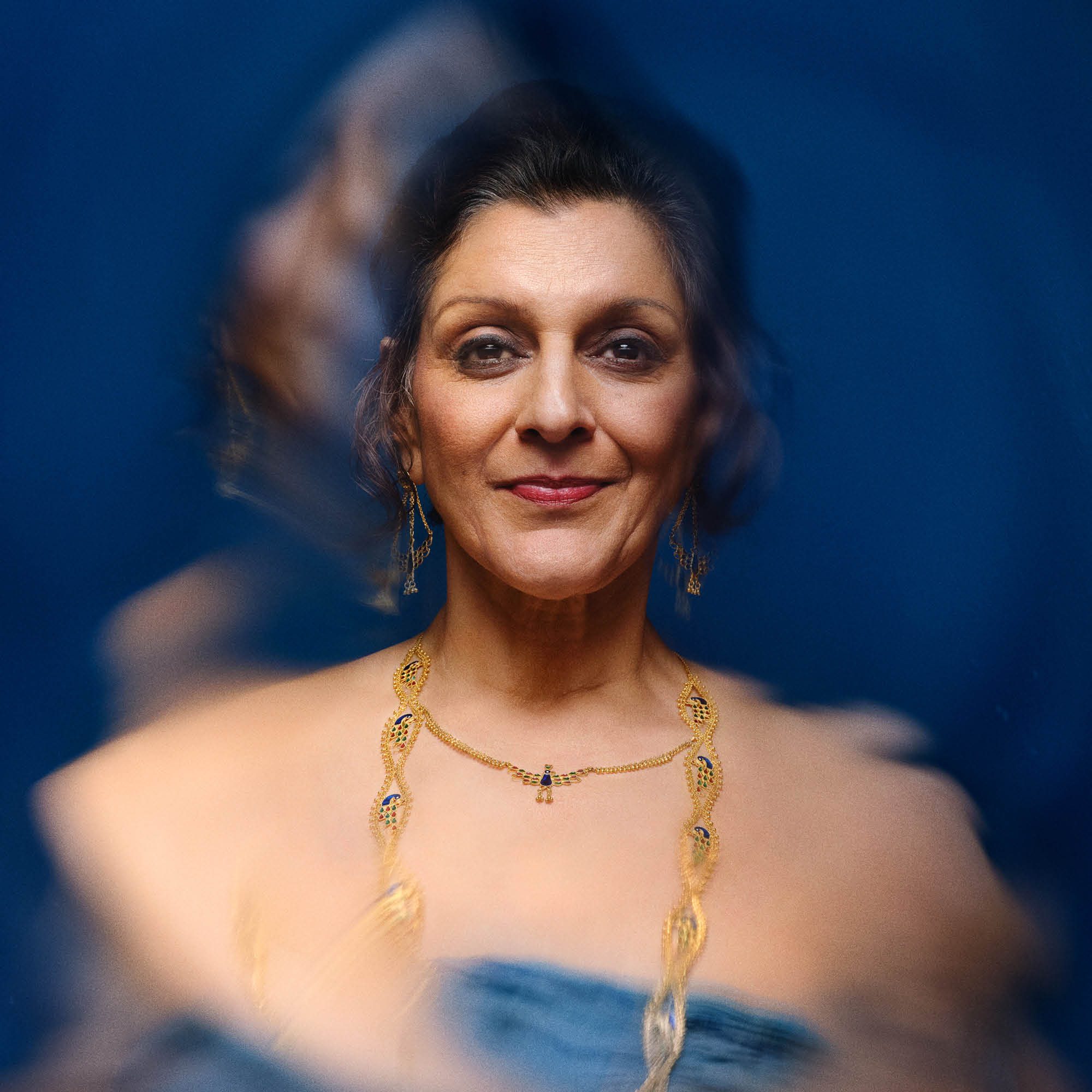 A blurred or shaken photo of a woman (Meera Syal), smiling and wearing gold jewellry and a blue dress.