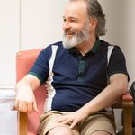 A man with a gray beard is sitting on a red chair, smiling and looking to his right. He is wearing a navy blue and white polo shirt and beige shorts. The background is a plain wall with a small portion of a table visible on the left.