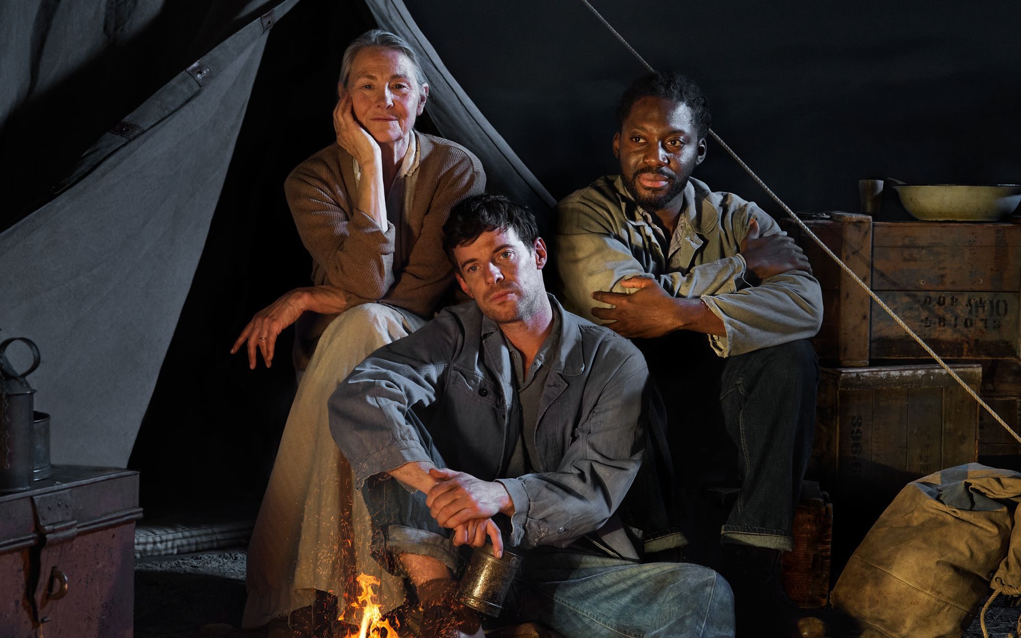 A woman and two men sit together in front of the open flap of a canvas tent. Packing boxes and canvas bags are either side of them and an open fire is before them.