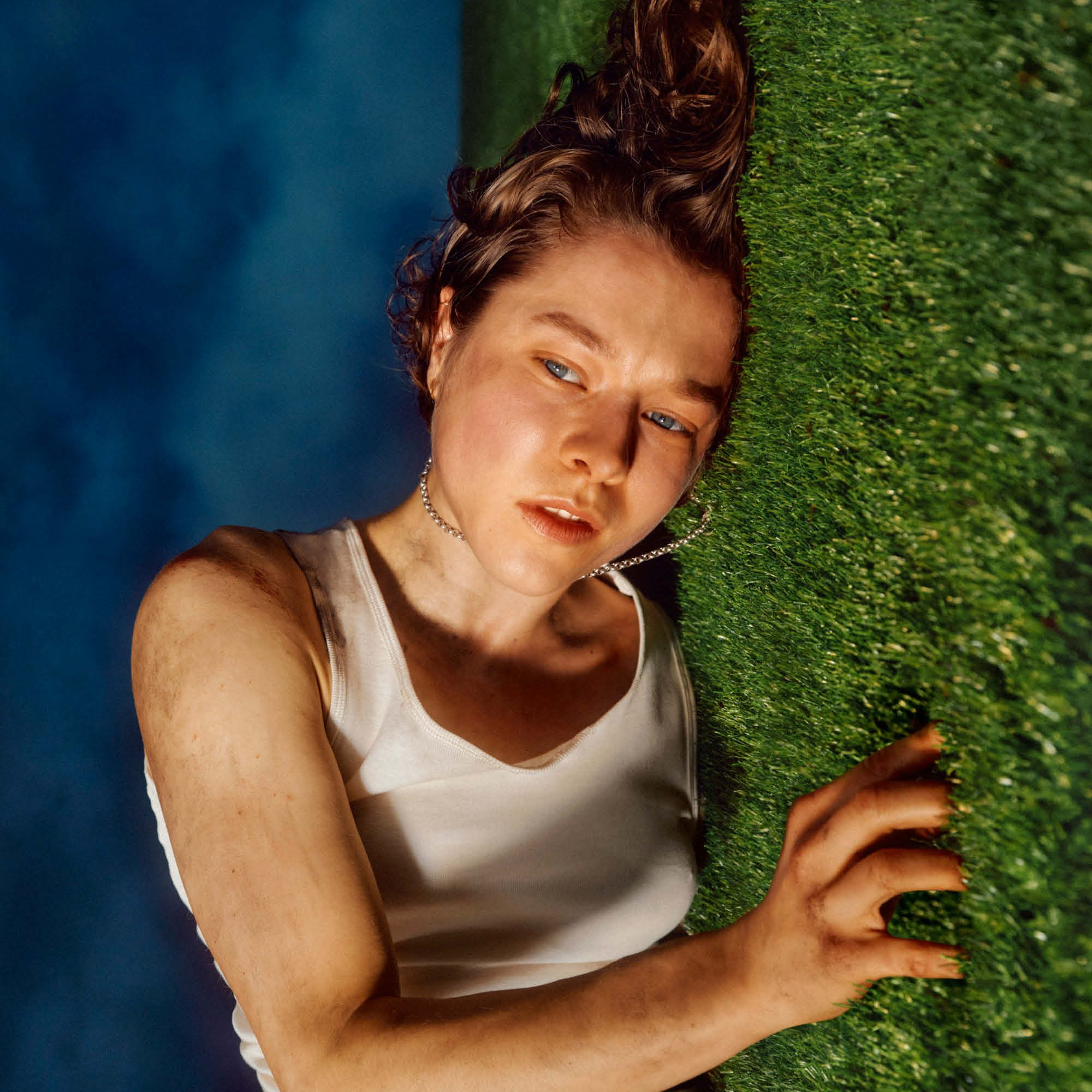 A person with short brown hair wearing a white tank top lies on their side on a grassy surface looking slightly upward. The image is taken from an angle that makes it appear as if the person is standing against a vertical grass wall, with a blue sky background.