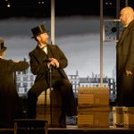 Three actors in 19th-century attire are on stage with a backdrop of historic buildings. One actor, seated on a stack of boxes and holding a cane, faces another standing actor. The third actor has his back turned to the camera. A clock is visible on the wall.