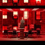 A person stands on a desk a stack of document boxes in front of him, in a futuristic, red-lit room with modern furniture, including desks and chairs. The setting has a dramatic, intense atmosphere with glowing red windows on the back wall.