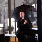 A person in a dark suit holds a black umbrella while sitting inside a modern room with large windows. There is a vase with flowers on a table nearby, and the room is dimly lit. The windows display reflections and outside scenery, hinting at an urban environment.