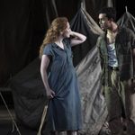 Two actors on stage, a woman with red hair wearing a blue dress, and a man with dark hair in a plaid shirt and brown trousers. They are standing in front of a backdrop of canvas tents. The woman touches her hair, while the man looks at her intently.