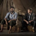 Two men, one barefoot and the other wearing boots, sit on wooden crates in front of a weathered tent. Both wear casual, rugged clothing, appearing contemplative. The man on the left gazes forward, while the man on the right looks at him.