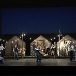 A lively stage scene set in front of three canvas tents with string lights. Performers dance, sing, and play instruments. The backdrop is a deep blue.