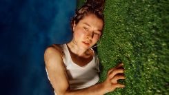 A person with short brown hair wearing a white tank top lies on their side on a grassy surface looking slightly upward. The image is taken from an angle that makes it appear as if the person is standing against a vertical grass wall, with a blue sky background.