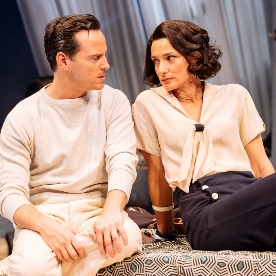 Two people sit closely together indoors, staring at each other with serious expressions. The man on the left wears a light gray sweatshirt, and the woman on the right wears a light blouse with a tie feature. Both have dark hair and are in a modern setting with soft lighting.