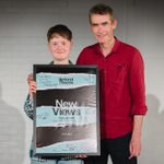 Two people stand and smile, holding a poster from the National Theatre's New Views Festival 2018. The person on the left is wearing a denim shirt, while the person on the right is wearing a red shirt. The poster is signed by multiple people and features event details.