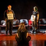 Three people are on stage in a theater. Two stand facing each other, holding signs reading 