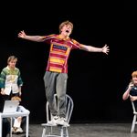 A young man stands on a chair with arms outstretched, energetically performing on stage while wearing a striped sports jersey. Around him, several people are engaged in various activities, such as typing on laptops, reading, or observing. The background is dimly lit.