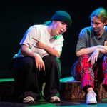 Two people on stage perform a dramatic scene. One, wearing a beanie and gesturing energetically, sits next to the other, who wears plaid trousers and listens attentively. A tote bag lies on the ground beside them. The background is dark, highlighting the performers.