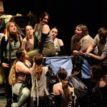 A group of young people in green and gray costumes perform on stage, gathered around a makeshift set piece made of trash bags and various items. They appear expressive and engaged, suggesting a dramatic scene. The background is dark, focusing attention on the performers.