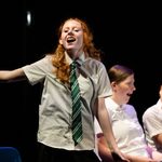 A young person with long red hair, wearing a white shirt and a green striped tie, sings passionately with one arm outstretched. In the background, two others in similar uniforms sit on blue chairs, one looking surprised and the other amused, against a dark backdrop.