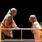 Two individuals with blonde hair stand on opposite sides of scaffolding poles. The person on the left wears a plaid shirt and leans forward, while the person on the right, in a light-colored top, also leans forward, engaging in an intense conversation against a dark background.