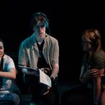 A group of young people sits on the floor in a dimly lit setting. They are engaged in a discussion. One person, wearing a headset and holding a notepad, looks directly at the camera, while another person points to the left. Blue stage lights are visible in the background.