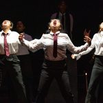 A group of people on stage, energetically performing. Three individuals wearing white shirts with red ties and dark trousers stand in front, their mouths open wide as if singing or shouting, with arms dramatically extended. Dim lighting creates a focused atmosphere.