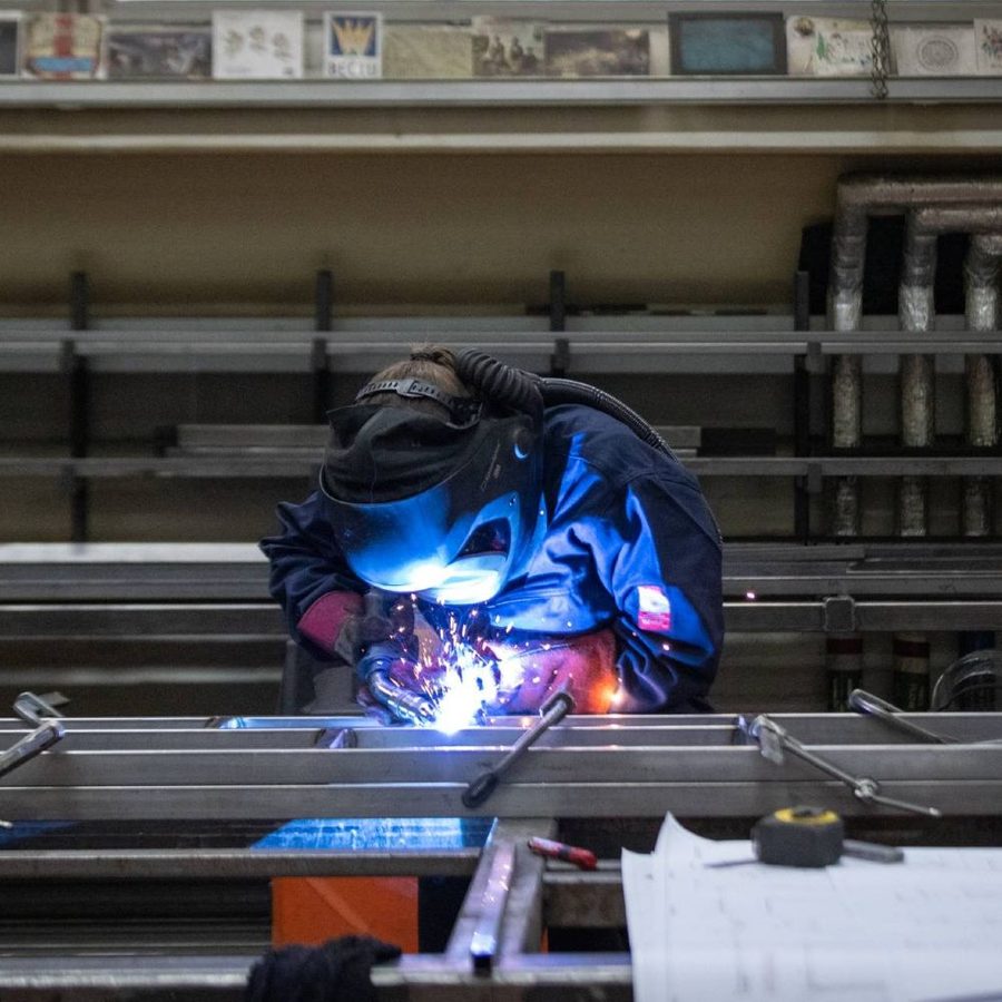 A person wearing protective gear and a welding helmet is performing welding work on a metal structure in a workshop. Various tools, pipes, and sheets are visible around the workspace.