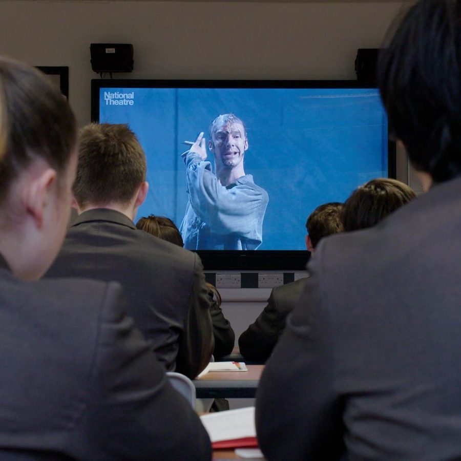 Students in a classroom watch a National Theatre production on a TV screen, featuring an actor in costume performing a scene. The students are seated with their backs to the camera, focused on the screen at the front of the room.