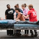 A group of six people, including a person lying on a bed, engage in a lively and friendly interaction. One person sits on the bed's edge, holding hands with the person lying down. They appear to be in a casual setting with a white brick wall in the background.