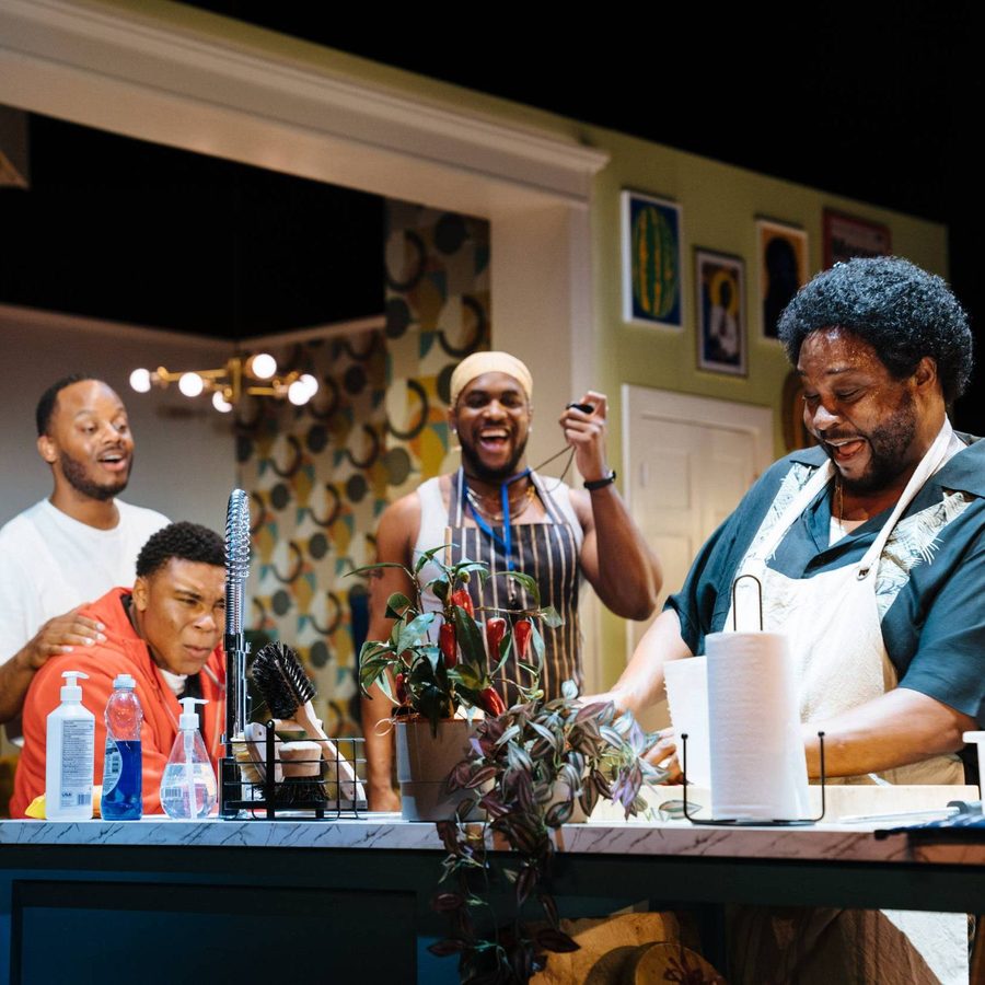 A joyous scene with five men in a kitchen. Four of them are standing together, smiling and laughing, while the fifth man stands at the stove with a smile. Various cleaning supplies and a plant are on the counter. The setting is bright and lively.