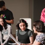 Four individuals are seated and holding scripts, engaged in what appears to be a theatrical rehearsal. One person in a pink hoodie sits at the back, while three others in the foreground are intently discussing their scripts.