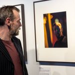 A man with a beard, wearing a striped blazer and red shirt, looks at a framed photo on a gallery wall. The photo depicts the same man backstage in a theatre and is dimly lit.