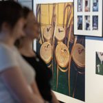 Two people view framed photographs in a photography exhibition. The main image on display shows several wooden chairs with woven seats, each holding a smoking pipe. Smaller photos are also visible next to the main picture.