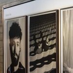 Three framed black and white photographs are displayed on a wall. The image on the left shows a portrait of a man with curly hair and a beard. The center features two people sat in theatre stalls seats. The right image depicts a person carrying boxes.