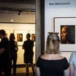 Several people are viewing photographs at an exhibition. The walls feature framed portraits on gallery boards with a white background. The room has a warm, ambient lighting, and visitors are engaged in looking at the artworks.