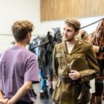 People in a rehearsal space: one person in a military uniform talking to another in a purple shirt, others in the background, and one person operating a large horse puppet. Scene conveys a theatrical preparation with focus on detailed military costumes and puppets.