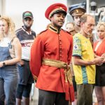 A group of people stand in a room, some holding musical instruments. One person in a red uniform and hat stands out in the center, looking forward with a serious expression. Others around him appear to be listening attentively or preparing for a performance.