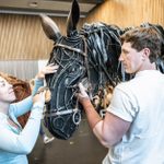 Two people handling a large horse puppet made of dark, metallic materials with intricate details. They are indoors with a wooden wall and other people in the background. The person on the left is touching the puppet's face, and the person on the right is holding its reins.