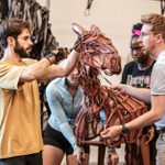 Four people are working together to manipulate a large, detailed horse puppet. The puppet's structure is made of natural-colored materials, including wood and leather. The background is to be an indoor studio with various equipment.