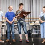 Four people are standing in a brightly lit room with a gray curtain background. Two men in casual blue tops and jeans listen attentively to a man with curly hair, holding a large open book. A woman in a light blue outfit also listens, holding a smaller book.