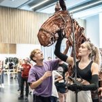 A group of people are in rehearsal in a spacious room. A woman handles a large, articulated puppet of a horse, while a man assists her. Others in the background observe and interact, suggesting a collaborative rehearsal environment.