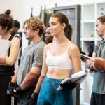 A group of people stand in a rehearsal room, most holding scripts or papers. They appear focused, with some wearing casual fitness attire and gloves. The setting suggests a practice session, for a performance.