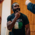 A man with a beard and short hair is standing indoors, wearing a black T-shirt with a Nigerian flag and the word 