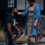 Two people are in the photo. One person, wearing a black t-shirt and gray pants, is kneeling. The other person, in a patterned dress and blue shawl, is standing and gently touching the kneeling person's face.