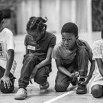 Four children kneel on the ground, each with focused expressions. The image is in black and white.