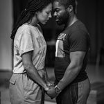 A black and white image depicts two people standing closely, holding hands and touching foreheads. Both appear to be in a serious, emotional moment. The person on the left wears a casual t-shirt and trousers, while the person on the right wears a t-shirt and shorts.