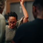 A woman wearing glasses and a textured grey top raises their right arm, appearing to be in the middle of a discussion. Two other partially visible individuals are in the foreground, slightly out of focus.