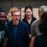 A group of people, including a man in glasses with a mustache, gather closely and appear to be engaged in a discussion. The background is blurred, highlighting the group's faces and attire.